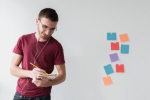 A man wearing headphones and glasses writing in a notebook at a desk, focused and studying, representing a modern professional building new skills.